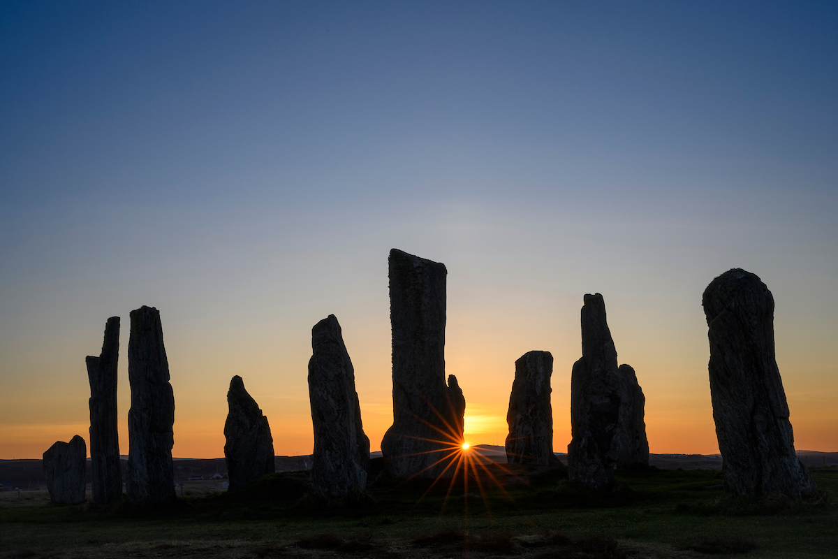 The Calanais Standing Stones silhouetted against a clear sky at sunrise, with a sunstar on the horizon between the stones